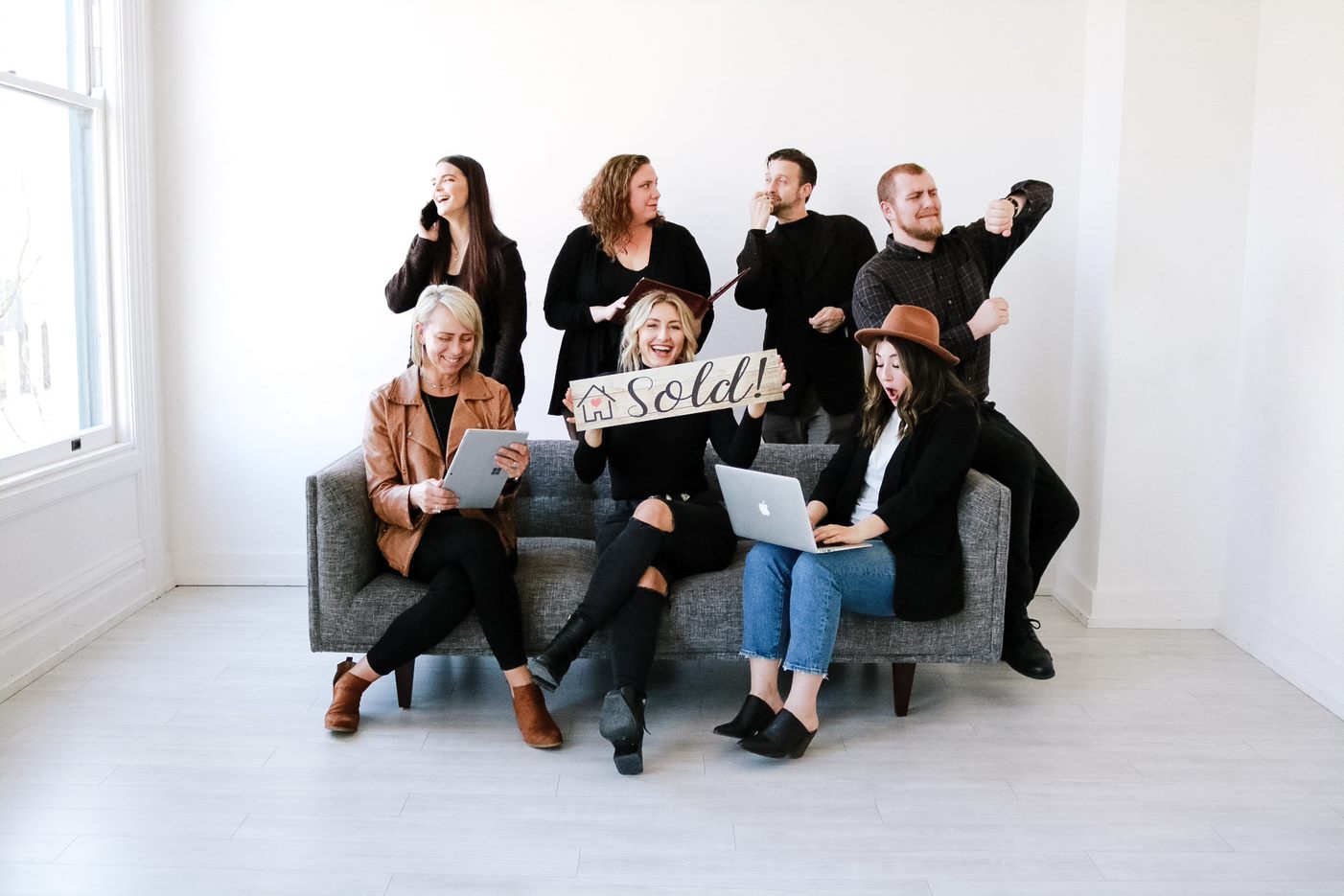 Group of people in business casual attire pretending to be doing various 'real estate agent' tasks (asnwering phones, on laptop, holding a 'sold' sign, etc) around a couch in an empty room.